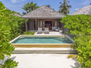 Luxusní plážová vila s&nbsp;vlastním bazénem, obklopená svěžími zelenými keři, lehátky a&nbsp;zastíněnou terasou pod dřevěnou střechou, s&nbsp;palmami a&nbsp;modrou oblohou v&nbsp;pozadí.