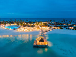 Letecký pohled na&nbsp;plážové letovisko za&nbsp;soumraku se svítícími řetězovými světly, bazénem, palmami, písečným pobřežím a&nbsp;dřevěným molem vedoucím k&nbsp;chatě se slaměnou střechou nad klidnou tyrkysovou vodou.