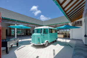 Tyrkysový kamion s&nbsp;jídlem parkuje na&nbsp;sluncem ozářeném otevřeném nádvoří, obklopeném modrými slunečníky, moderními budovami, venkovním posezením a&nbsp;stolním fotbalem pod částečně zamračenou oblohou.