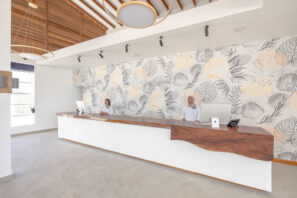 Moderní hotelová recepce se dvěma recepčními za&nbsp;dlouhým bílým a&nbsp;dřevěným stolem, světlými stěnami, velkými okny a&nbsp;ozdobnou stěnou s&nbsp;listovým vzorem.