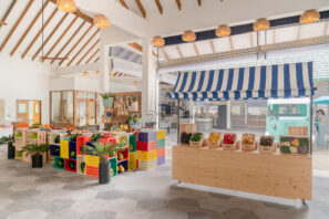 Jasně osvětlený krytý trh s&nbsp;barevnými bednami s&nbsp;čerstvým ovocem a&nbsp;zeleninou, dřevěným stánkem s&nbsp;produkty a&nbsp;pruhovanou modrobílou stříškou. Prostor má vysoké stropy a&nbsp;velká okna.