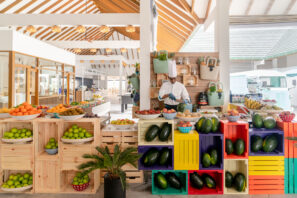 Kuchař v&nbsp;bílé uniformě připravuje jídlo za&nbsp;barevným stánkem s&nbsp;ovocem plným pomerančů, jablek, limetek a&nbsp;avokád ve&nbsp;světlém, moderním krytém marketu s&nbsp;dřevěnými trámy a&nbsp;přirozeným světlem.