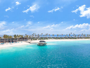 Tropická pláž s&nbsp;průzračnou tyrkysovou vodou, bílým pískem a&nbsp;palmami. Nad vodou se táhne dřevěné molo s&nbsp;malou chatkou ve&nbsp;stínu. V&nbsp;pozadí jsou pod jasně modrou oblohou vidět budovy a&nbsp;další palmy.