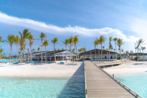 Dřevěné molo vede na&nbsp;bílou písečnou pláž lemovanou palmami a&nbsp;dvěma moderními budovami, v&nbsp;popředí je průzračná tyrkysová voda a&nbsp;nad hlavou modrá obloha.