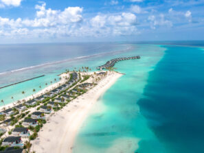 Letecký pohled na&nbsp;tropický ostrovní resort s&nbsp;bungalovy nad vodou, bílými písečnými plážemi, tyrkysovým oceánem, palmami a&nbsp;jasně modrou oblohou s&nbsp;rozptýlenými mraky.