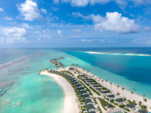 Letecký pohled na&nbsp;tropický ostrov s&nbsp;bílými písečnými plážemi, tyrkysovou vodou, řadami vil nad vodou a&nbsp;na pláži, palmami a&nbsp;malými vzdálenými písečnými přesypy pod modrou oblohou s&nbsp;rozptýlenými mraky.