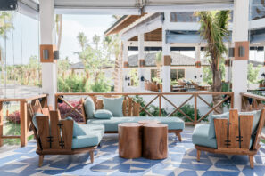 Moderní venkovní lounge s&nbsp;modrými dřevěnými židlemi s&nbsp;polštáři, odpovídajícím loveseatem a&nbsp;dvěma kulatými dřevěnými stoly na&nbsp;geometrické modrobílé dlažbě, obklopené tropickými rostlinami a&nbsp;slunečním světlem.