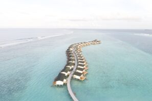 Nad průzračně tyrkysovou vodou oceánu se táhne řada nadmořských vil propojených zakřivenou dřevěnou promenádou, obklopených klidným, rozlehlým mořem pod lehce zataženou oblohou.