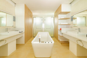 Moderní prostorná koupelna s&nbsp;centrální bílou volně stojící vanou, dvěma umyvadly se zrcadly na&nbsp;každé straně, skříňkami ze světlého dřeva, otevřenými policemi s&nbsp;ručníky a&nbsp;dveřmi z&nbsp;matného skla v&nbsp;pozadí.