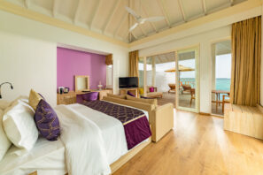 Světlý, moderní hotelový pokoj s&nbsp;velkou postelí, fialovými akcenty, dřevěnou podlahou a&nbsp;posuvnými skleněnými dveřmi vedoucími na&nbsp;terasu s&nbsp;venkovním posezením a&nbsp;výhledem na&nbsp;oceán.