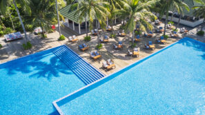 Letecký pohled na&nbsp;velký průzračně modrý bazén obklopený palmami a&nbsp;venkovním posezením se stoly a&nbsp;židlemi u&nbsp;pavilonu za&nbsp;slunečného dne.