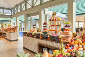 Barevný bufet s&nbsp;rozmanitým čerstvým ovocem a&nbsp;dezerty naaranžovanými na&nbsp;stupňovitých stojanech, dřevěných bednách a&nbsp;deskách ve světlé otevřené jídelně s&nbsp;velkými okny a&nbsp;zelenými střechami, které jsou vidět venku.