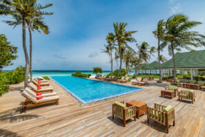 Z luxusního venkovního bazénu s&nbsp;lehátky, polstrovanými křesly a&nbsp;palmami je výhled na&nbsp;tyrkysový oceán pod modrou oblohou. Vpravo od dřevěné terasy se nachází krytý pavilon.
