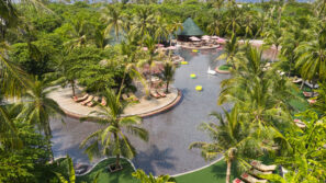 Letecký pohled na&nbsp;velký bazén v&nbsp;tropickém letovisku obklopený palmami, lehátky a&nbsp;slunečníky, s&nbsp;barevnými plováky ve vodě a&nbsp;bujnou zelení všude kolem.