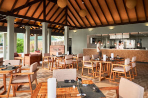 Prostorná moderní restaurace s&nbsp;dřevěnými stoly a&nbsp;polstrovanými židlemi, otevřenou kuchyní a&nbsp;vysokým stropem s&nbsp;odhalenými trámy. Kolem prochází člověk, zatímco v&nbsp;pozadí za&nbsp;pultem pracují kuchaři.