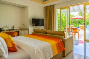 Světlý hotelový pokoj s&nbsp;postelí pokrytou oranžovým a&nbsp;žlutým povlečením, televizí, malým pracovním prostorem a&nbsp;posuvnými dveřmi, které se otevírají na&nbsp;terasu s&nbsp;židlemi, stolem a&nbsp;červeným slunečníkem u&nbsp;bazénu.