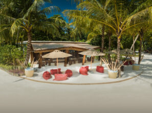 Tropický plážový bar s&nbsp;dřevěným nábytkem, palmami, béžovými slunečníky a&nbsp;barevnými pytli s&nbsp;fazolemi na&nbsp;bílém písku, obklopený bujnou zelení pod jasně modrou oblohou.