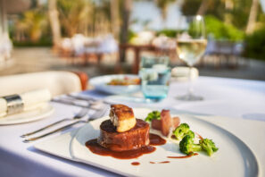 Na prostřeném jídelním stole s&nbsp;vínem a&nbsp;skleněným nádobím pod širým nebem se podává gurmánský pokrm - steak s&nbsp;foie gras, podávaný s&nbsp;omáčkou, brokolicí a&nbsp;zeleninou na&nbsp;bílém talíři.