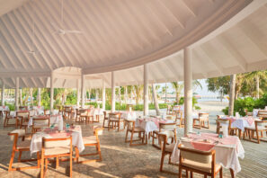 Restaurace pod širým nebem na&nbsp;pláži s&nbsp;bílými sloupy a&nbsp;stropem, úhledně prostřenými stoly s&nbsp;růžovými ubrusy a&nbsp;židlemi, s&nbsp;výhledem na&nbsp;palmy a&nbsp;písek, do&nbsp;kterého proudí sluneční světlo.