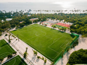 Letecký pohled na&nbsp;svěží zelené fotbalové hřiště obklopené stromy, v&nbsp;jehož blízkosti se nacházejí další sportovní hřiště. V&nbsp;pozadí jsou pod jasnou oblohou vidět palmy, budovy a&nbsp;oceán.