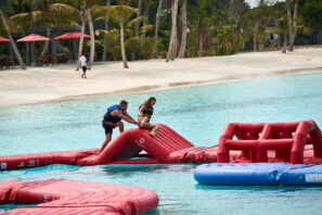 Dva lidé v&nbsp;záchranných vestách si hrají na&nbsp;červené nafukovací překážkové dráze ve vodě poblíž tropické pláže s&nbsp;palmami, pískem a&nbsp;červenými slunečníky v&nbsp;pozadí.