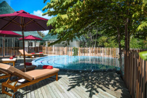 Malý kulatý venkovní bazén je obklopen dřevěnou terasou s&nbsp;lehátky a&nbsp;červenými slunečníky. Stromy poskytují stín a&nbsp;areál je ohraničen dřevěným plotem. V&nbsp;pozadí je vidět modrá obloha a&nbsp;zeleň.