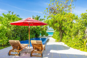 Dvě dřevěná lehátka s&nbsp;červeným slunečníkem stojí vedle malého bazénu obklopeného svěží zelení. Bazén vede k&nbsp;výhledu na&nbsp;oceán pod jasně modrou oblohou.