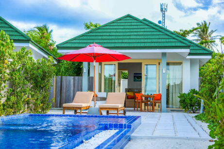 Moderní vila se zelenou střechou a&nbsp;prosklenými dveřmi se dvěma lehátky pod červeným slunečníkem u&nbsp;modrého bazénu obklopeného bujnou zelení.