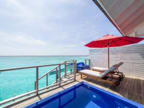 Dvě lehátka pod červeným slunečníkem na&nbsp;terase vedle malého bazénu s&nbsp;výhledem na&nbsp;průzračně tyrkysovou vodu oceánu pod slunečnou oblohou.