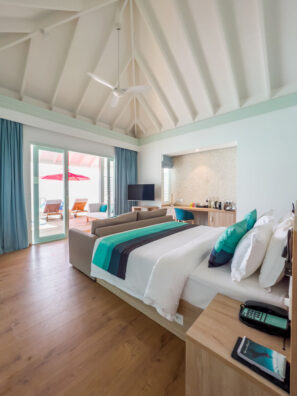 Moderní, vzdušný hotelový pokoj s&nbsp;úhledně ustlanou postelí, čajovými a&nbsp;černými akcenty, posezením, dřevěnou podlahou a&nbsp;velkými prosklenými dveřmi na&nbsp;sluncem ozářenou terasu s&nbsp;lehátky a&nbsp;červeným slunečníkem.