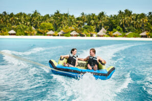 Dva lidé v&nbsp;záchranných vestách jedou společně na&nbsp;nafukovacím člunu, který je tažen po&nbsp;průzračně modré vodě, v&nbsp;pozadí jsou vidět tropické palmy a&nbsp;plážové chatky.