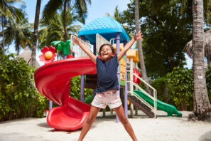 Šťastné dítě v&nbsp;šortkách a&nbsp;tričku skáče se zdviženýma rukama před barevnou skluzavkou na&nbsp;dětském hřišti obklopeném palmami a&nbsp;zelení za&nbsp;slunečného dne.