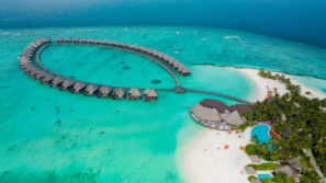 Letecký pohled na&nbsp;tropický resort s&nbsp;bungalovy nad vodou uspořádanými do&nbsp;půlkruhu nad tyrkysovou vodou, bílou písečnou pláží, palmami a&nbsp;bazénem obklopeným zelení.