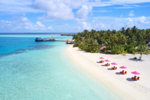 Tropická pláž s&nbsp;průzračnou tyrkysovou vodou, bílým pískem, červenými lehátky s&nbsp;růžovými slunečníky, bujnými palmami a&nbsp;přístavištěm s&nbsp;loďkami pod jasně modrou oblohou s&nbsp;roztroušenými mraky.