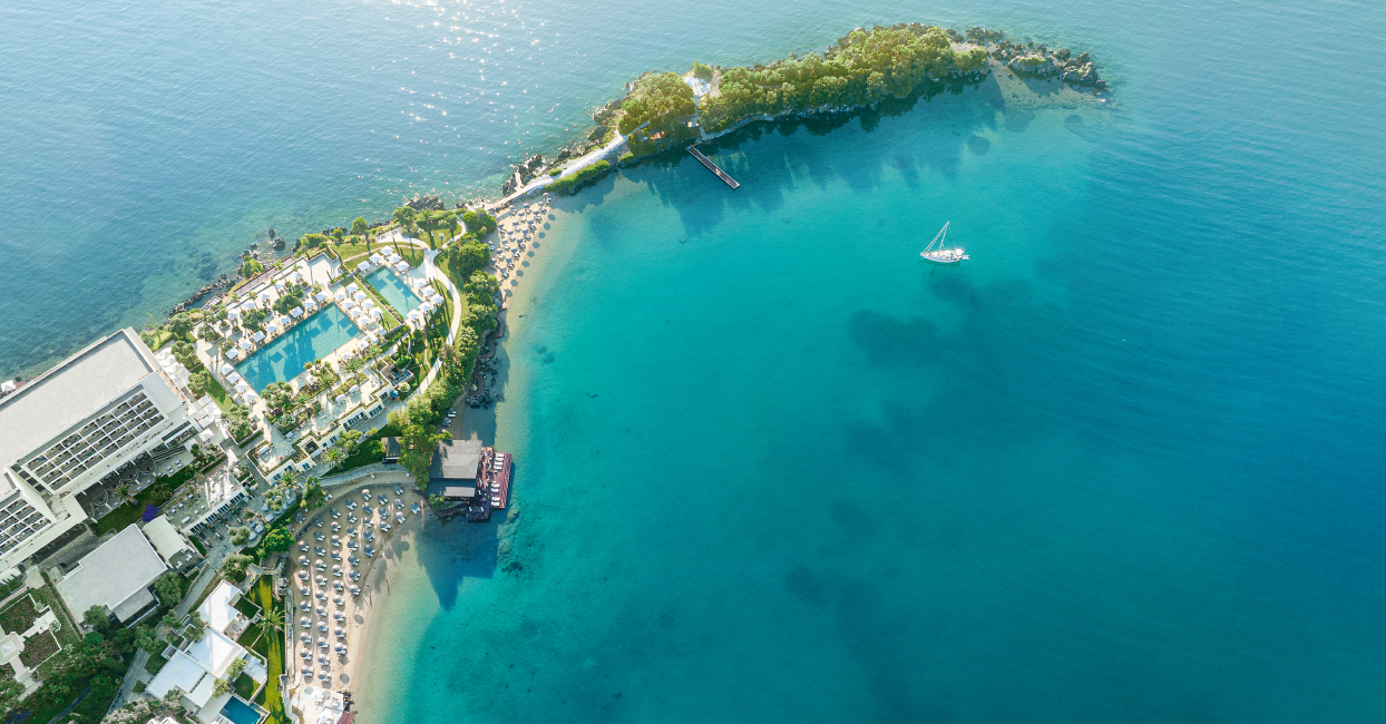 Letecký pohled na&nbsp;letovisko s&nbsp;bazény, lehátky a&nbsp;slunečníky u&nbsp;průzračně modré vody; nedaleko pluje plachetnice a&nbsp;malý poloostrov pokrývá zeleň.