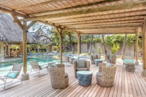 Venkovní odpočívárna s proutěnými židlemi a stoly na dřevěné terase pod pergolou s výhledem na průzračný bazén, obklopená tropickými rostlinami a budovami s doškovou střechou.