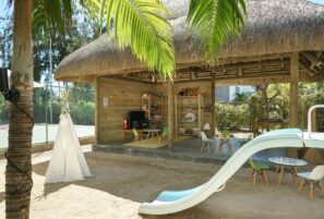 Písečný venkovní dětský koutek s modrou skluzavkou, bílým teepee a malými stolky a židlemi, vedle dřevěné chatky pod širým nebem s policemi, hračkami a televizí, obklopený bujnou zelení a palmami.