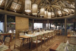 Restaurace s tropickou tématikou, dřevěnými stoly a židlemi, doškovou střechou, měkkými závěsnými světly a úhledně prostřenými stoly, které vytvářejí příjemnou atmosféru.