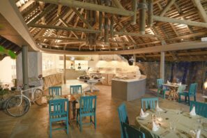 Rustikální restaurace pod širým nebem s tyrkysovými dřevěnými židlemi, kulatými stoly k stolování, bufetem, kamenným barem a dřevěnými trámy podpírajícími doškovou střechu; prostor zdobí bílé jízdní kolo.