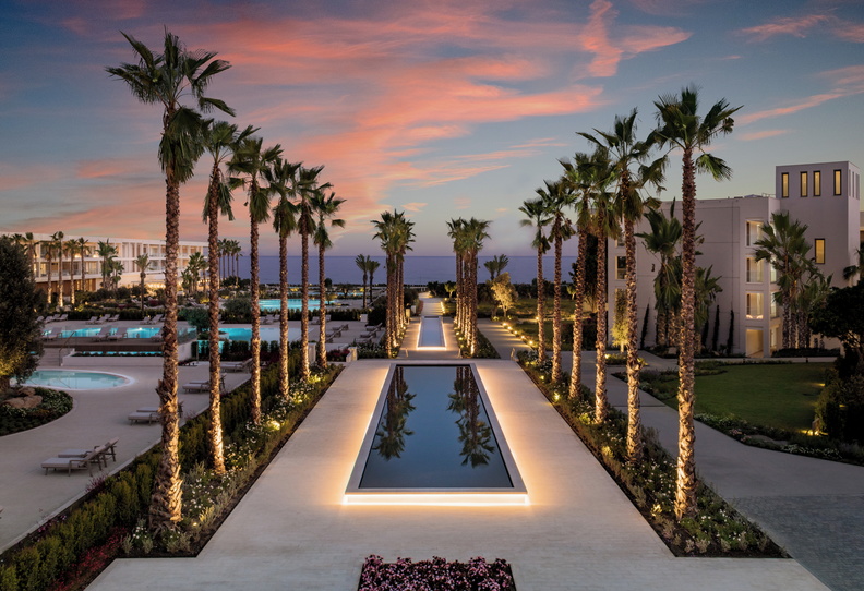 Široká cesta lemovaná vysokými palmami a&nbsp;dlouhým osvětleným zrcadlícím se bazénem vede při západu slunce směrem k&nbsp;oceánu a&nbsp;po obou stranách jsou vidět budovy resortu a&nbsp;bazény.