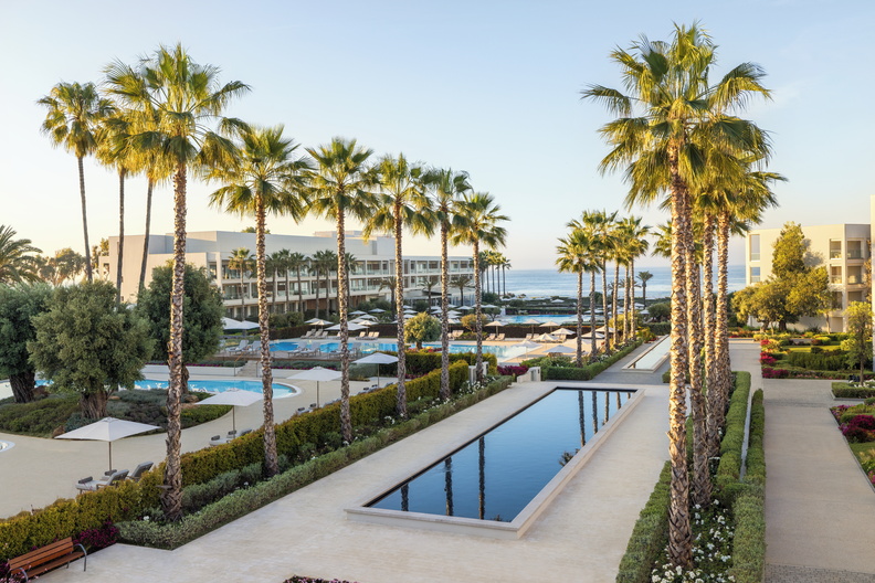Luxusní letovisko s&nbsp;palmami, zahradami, dlouhým reflexním bazénem, lehátky a&nbsp;bazény, s&nbsp;moderními budovami a&nbsp;oceánem viditelným v&nbsp;pozadí pod jasnou oblohou.