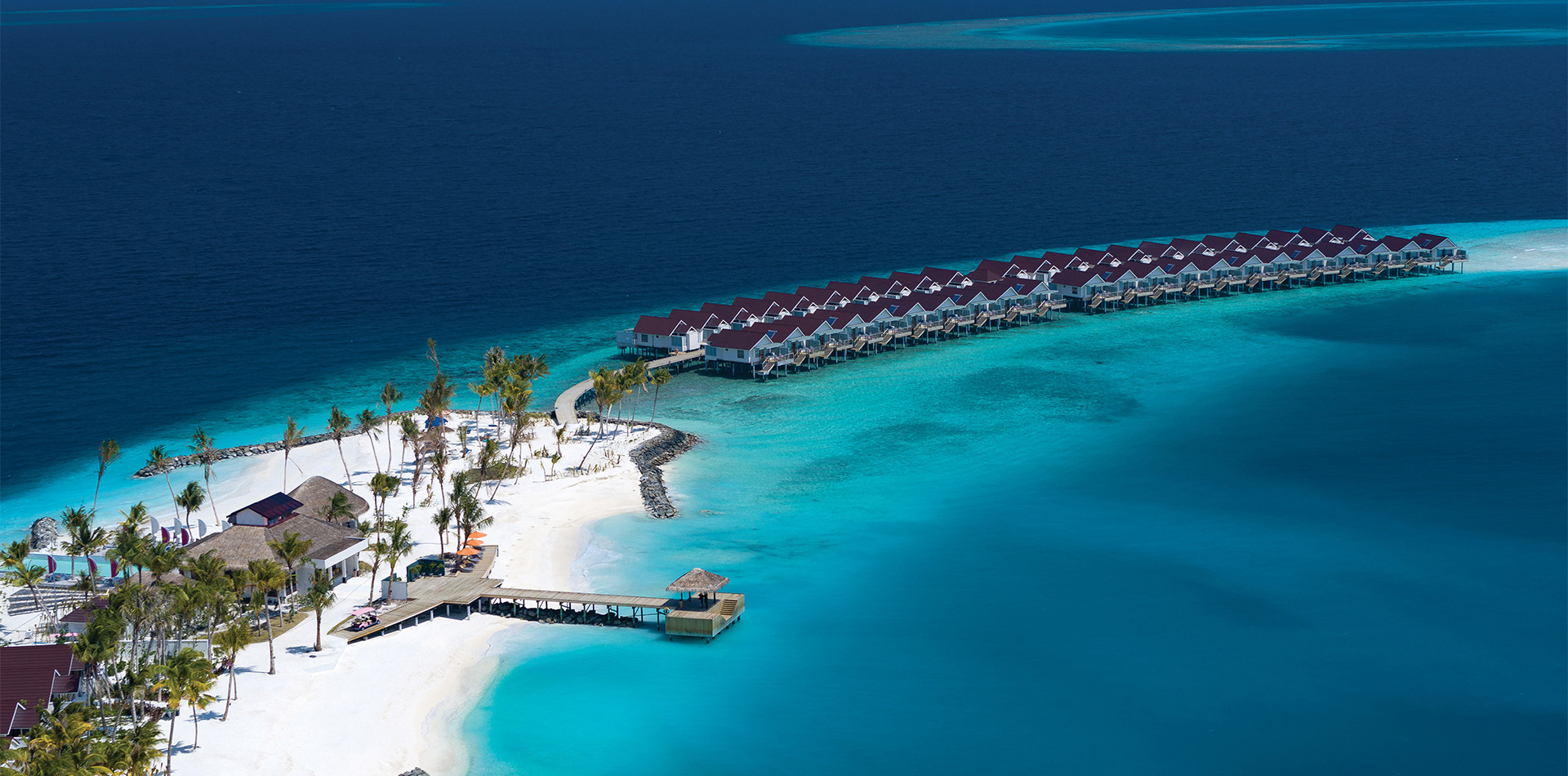 Letecký pohled na tropický ostrovní resort s vilami nad vodou, bílou písečnou pláží, palmami a průzračným tyrkysovým oceánem obklopeným hlubokou modrou vodou.