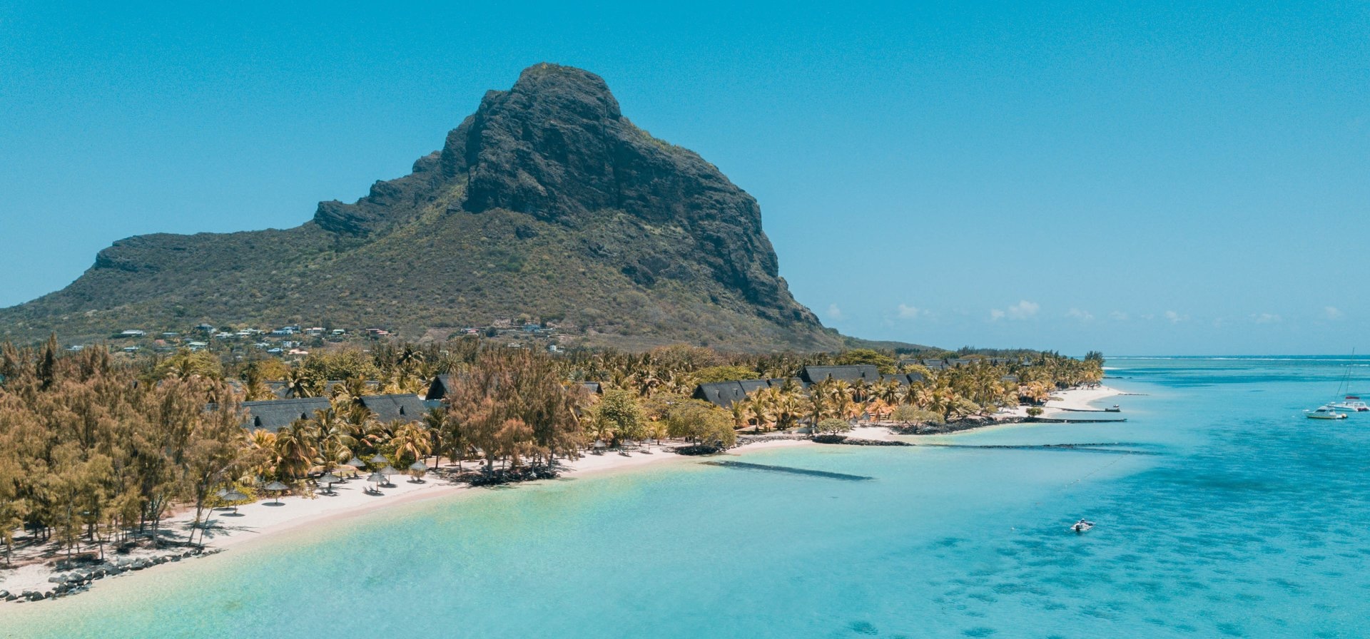 Letecký pohled na&nbsp;tropickou pláž s&nbsp;průzračnou tyrkysovou vodou, bílým pískem, palmami a&nbsp;výraznou horou tyčící se v&nbsp;pozadí pod jasně modrou oblohou.
