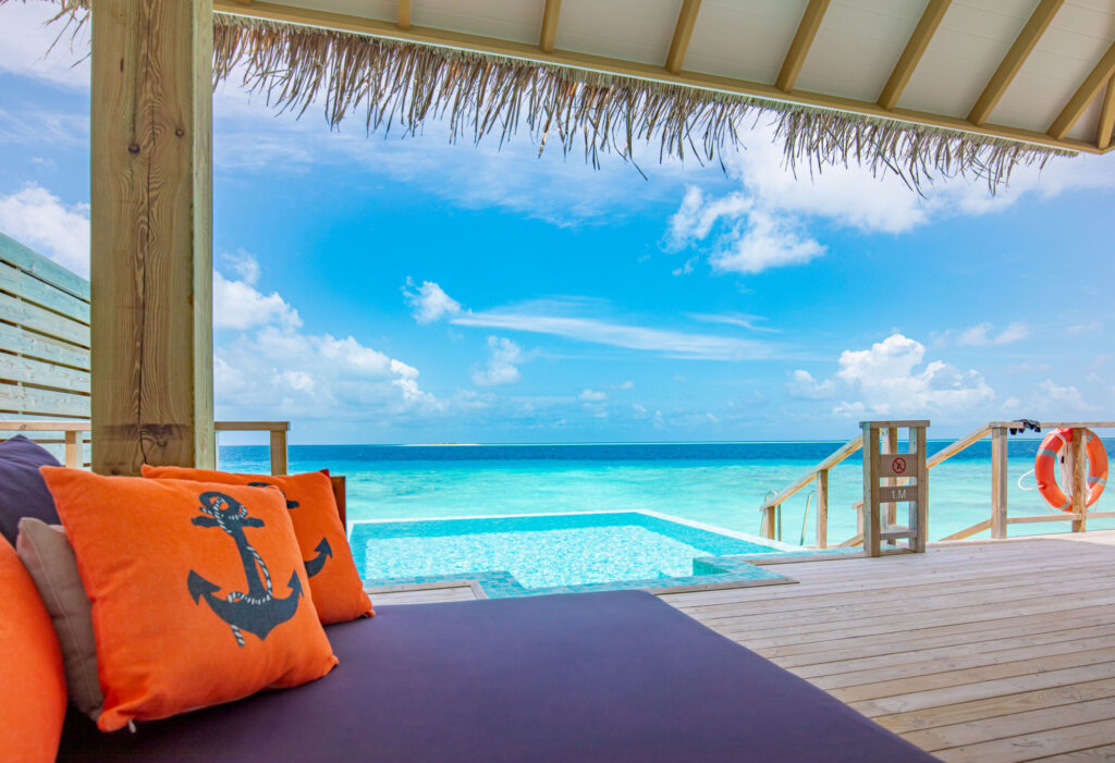 Pohled z&nbsp;terasy vodní vily v&nbsp;Iru Veli s&nbsp;infinity bazénem, tyrkysovým oceánem a&nbsp;stylovými oranžovými polštáři s&nbsp;kotvou.