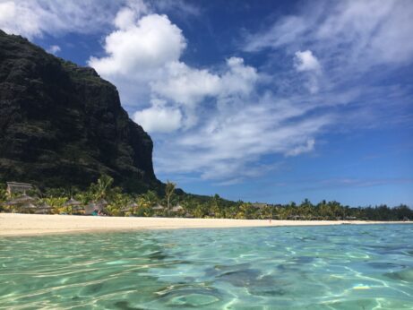 Průzračná tyrkysová voda v popředí, písečná pláž lemovaná palmami a slaměnými slunečníky v hotelu LUX* Le Morne a vysoký skalní útes vlevo, to vše pod jasně modrou oblohou s rozptýlenými mraky.