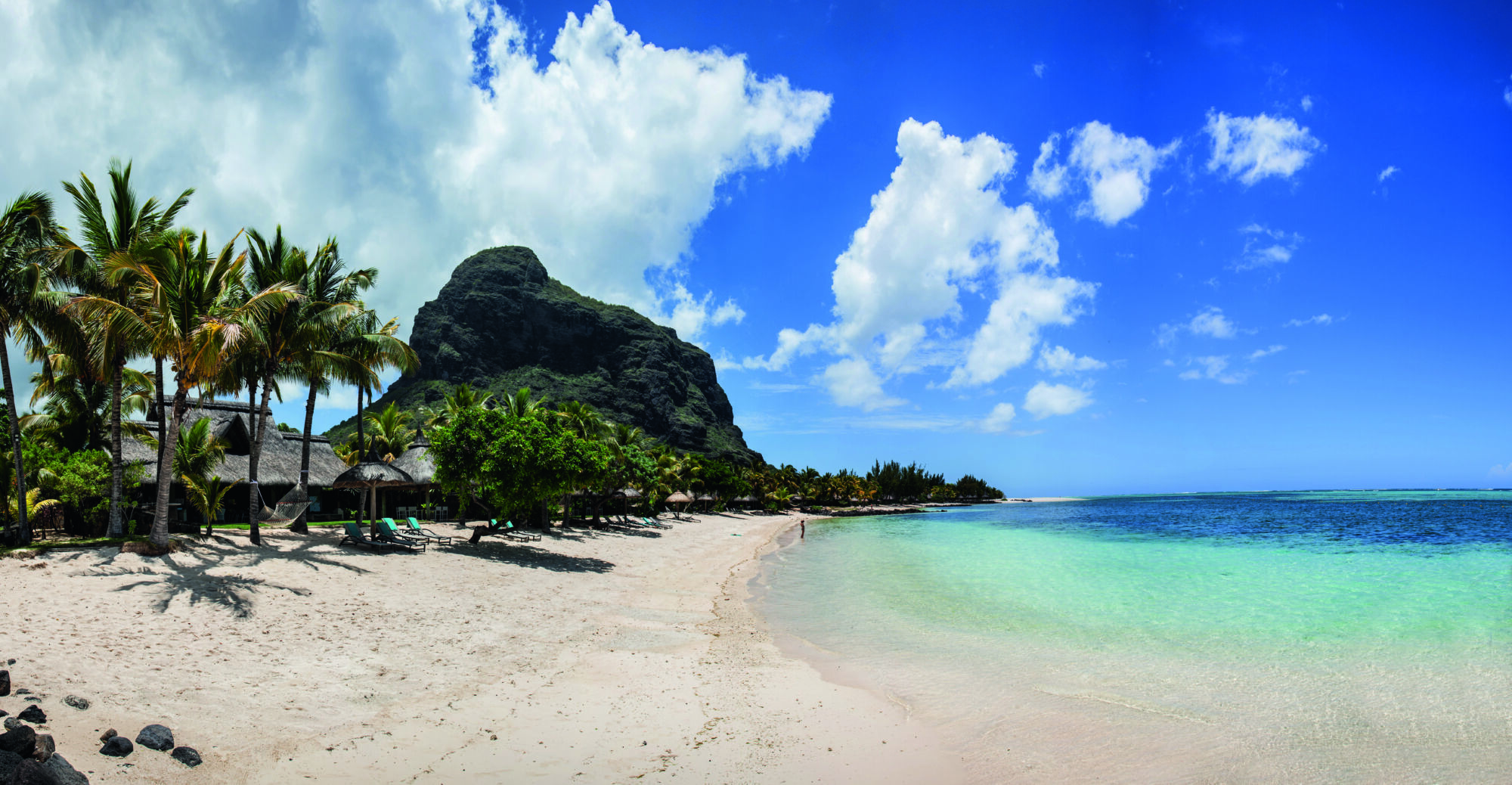 Tropická pláž s&nbsp;bílým pískem, průzračnou tyrkysovou vodou, palmami a&nbsp;chatkami s&nbsp;doškovými střechami. V&nbsp;pozadí se pod částečně zataženou modrou oblohou nedaleko luxusního 5* resortu Paradis Beachcomber tyčí úchvatná hora Le Morne.
