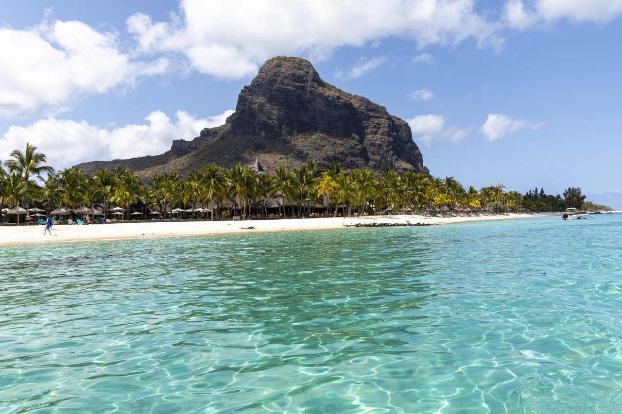 V popředí průzračná tyrkysová voda a&nbsp;písečná pláž lemovaná palmami. V&nbsp;pozadí se pod modrou oblohou s&nbsp;rozptýlenými mraky majestátně tyčí Le Morne - ikonický výhled nedaleko 5* resortu Paradis Beachcomber.