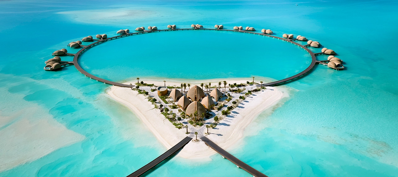 Letecký pohled na&nbsp;tropický resort s&nbsp;nadmořskými vilami v&nbsp;kruhovém uspořádání nad tyrkysovou vodou, ideální pro&nbsp;luxusní cestování. Dřevěné chodníčky navazují na&nbsp;bílý písečný ostrov s&nbsp;moderními budovami a&nbsp;bujnou zelení, připomínající útěky v&nbsp;Saudské Arábii.