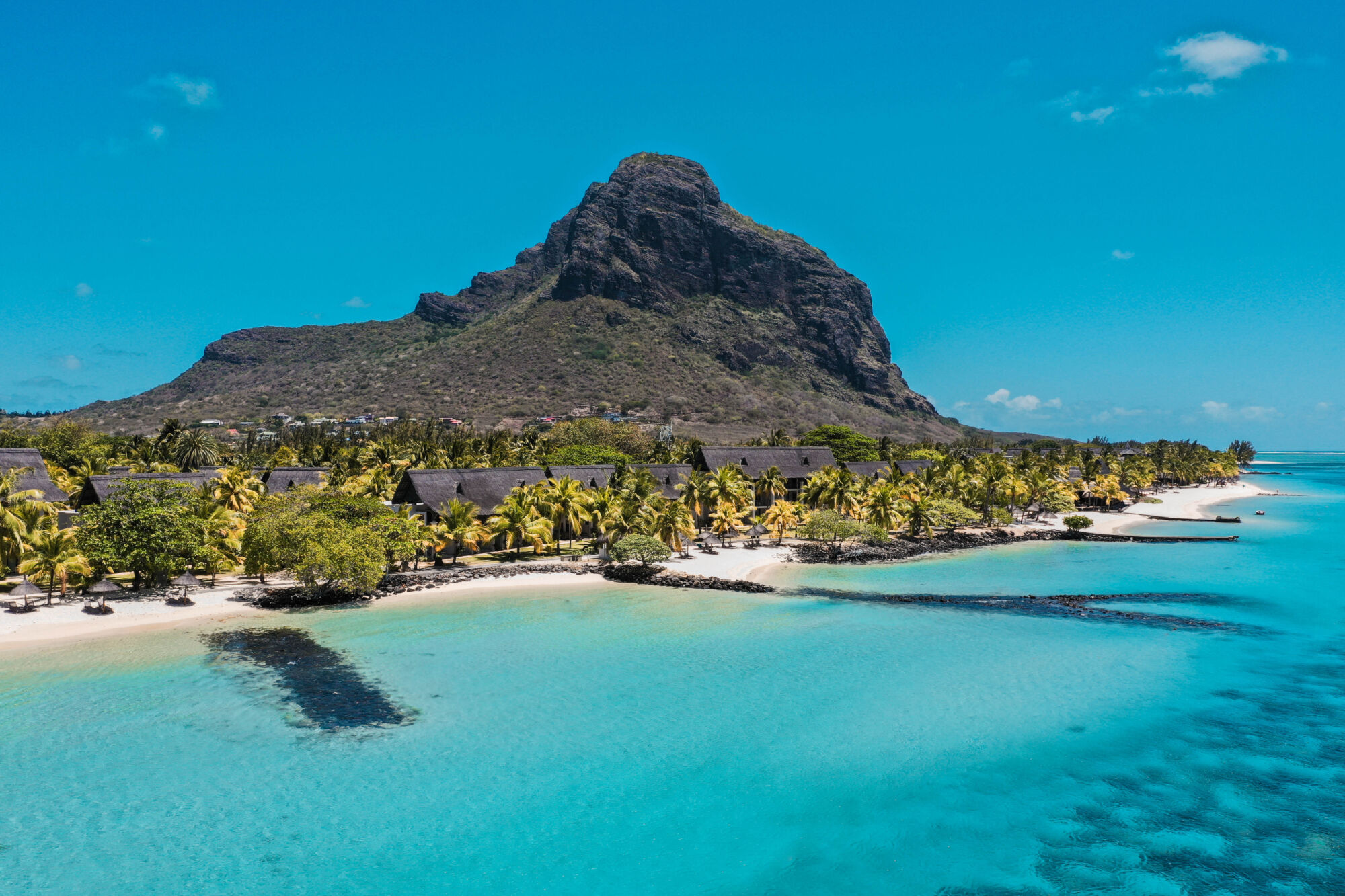 Tyrkysová laguna s&nbsp;bílými písečnými plážemi lemovanými palmami, tropickými chatkami a&nbsp;bujnou zelení. V&nbsp;pozadí se pod jasně modrou oblohou zvedá velká skalnatá hora.