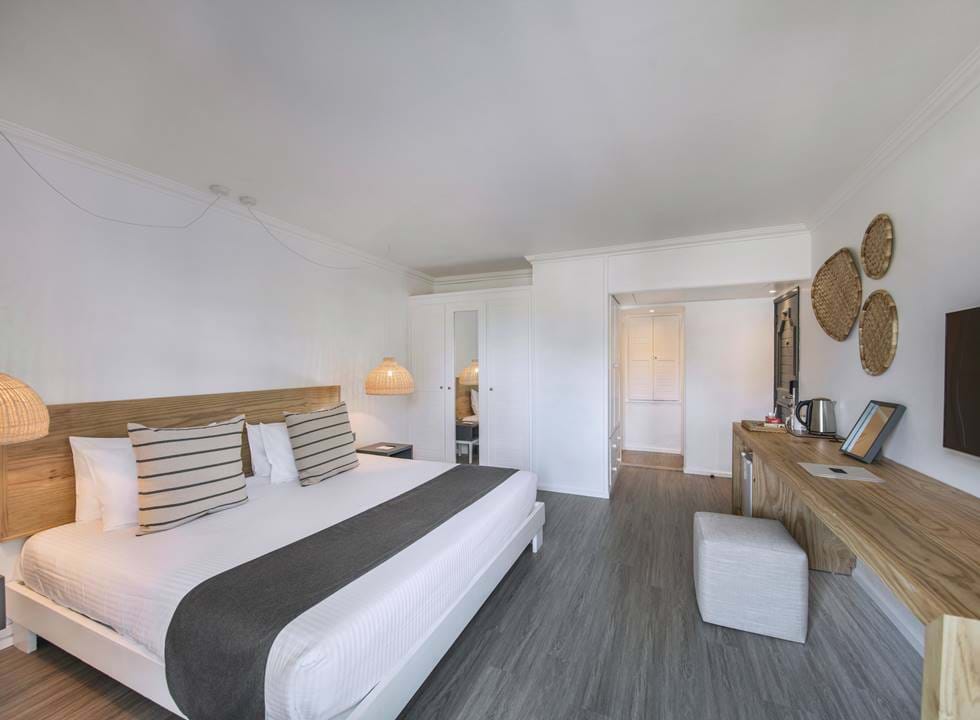 Moderní hotelový pokoj s velkou postelí, pruhovanými polštáři, dřevěným čelem postele, proutěnými lampami, psacím stolem s rychlovarnou konvicí a šálky, nástěnnou televizí a tkanými koši zdobícími bílou stěnu.